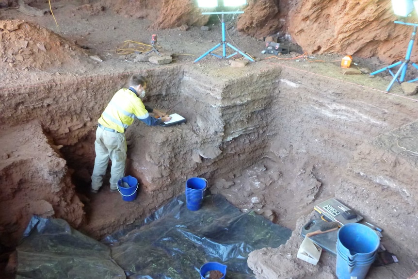 A man inside a dig pit makes notes as he excavates artefacts.