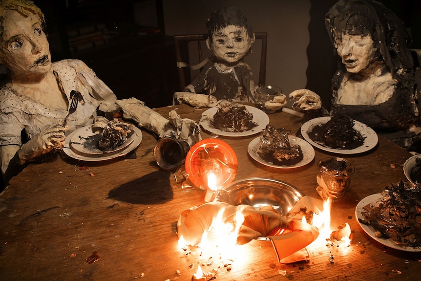 Colour image from 2018 stop-motion film La Casa Lobo of 3 painted puppets made of cardboard sitting around a dining table.
