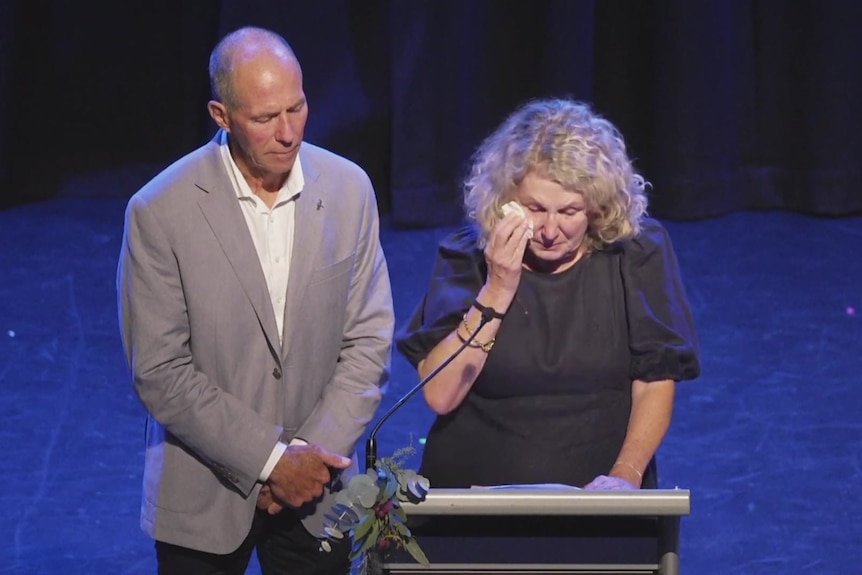 A tearful couple speak at a podium in dark clothes, Older, mid fifties
