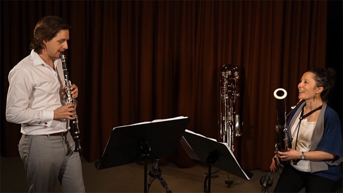 A man in a white shirt and grey trousers plays the clarinet while a woman holding a bassoon looks on, smiling at him.