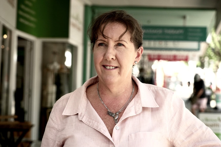 A dark-haired woman in a pale pink button-up shirt, stands in front of a row of shops