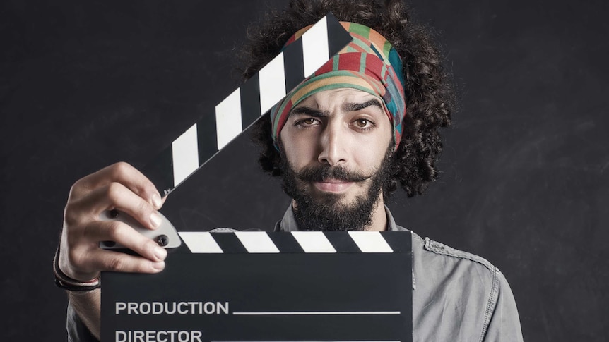 A man with a beard and a scarf on his head holding a film clapper board