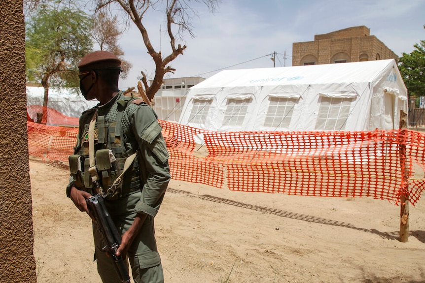 A soldier stands with a gun in front of a white tent.