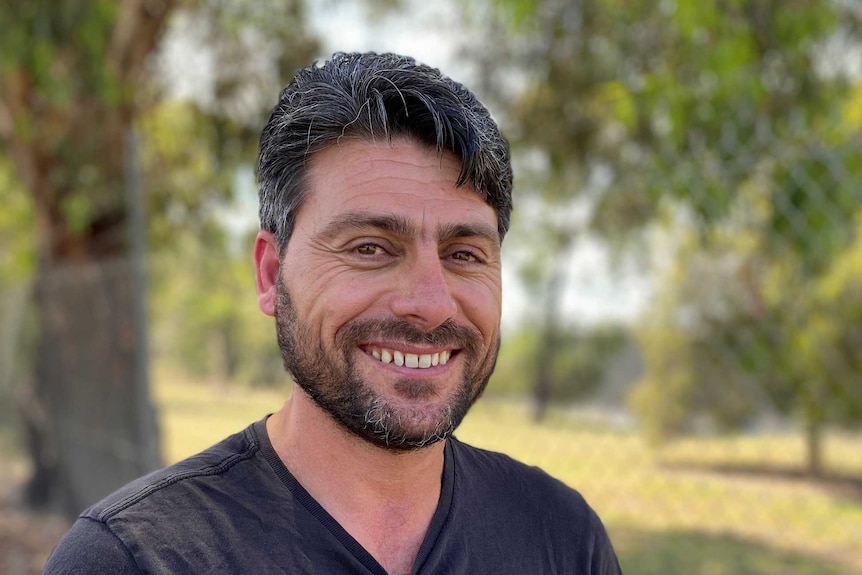 A smiling man with dark hair and a pair of sunglasses in his t-shirt.