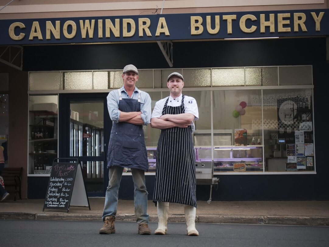 Two men in butcher outfits standing in front of a butcher shop