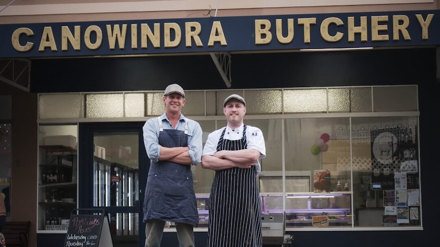 Two men in butcher outfits standing in front of a butcher shop