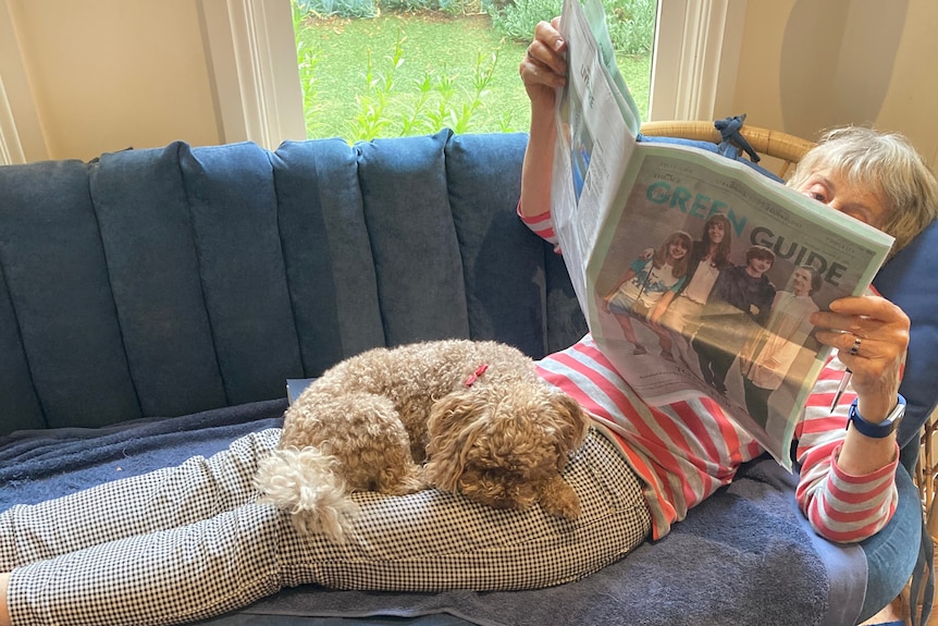 Katriona reads a newspaper while her dog Fudge sits on her lap.