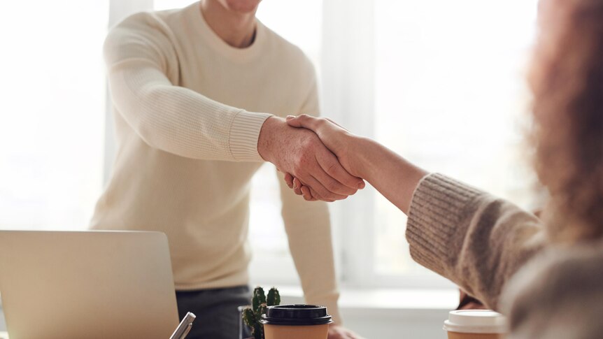 man in cream jumper standing at a desk shaking hands with a woman who is sitting down. 