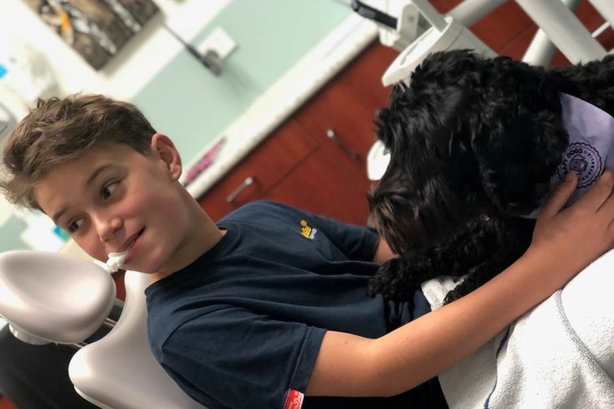 Alex Gorman holds dental therapy dog Comet on his lap while sitting in a dental chair.