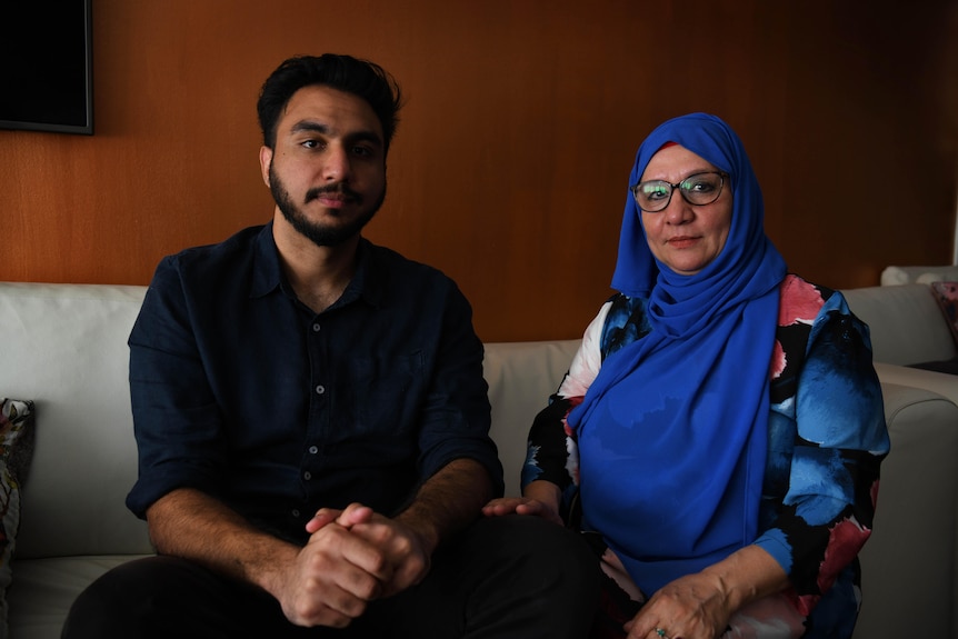 A man with dark hair and trimmed beard sits next to his mother who wears glasses and neon blue headscarf