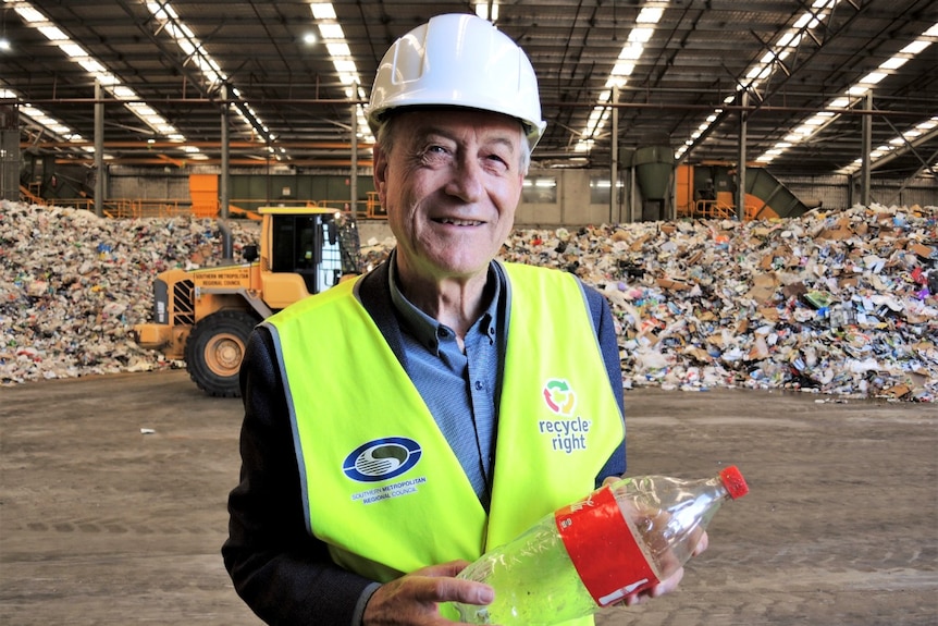 A man wearing a yellow vest and white hard hat holds a plastic drink bottle at a recycling facility.