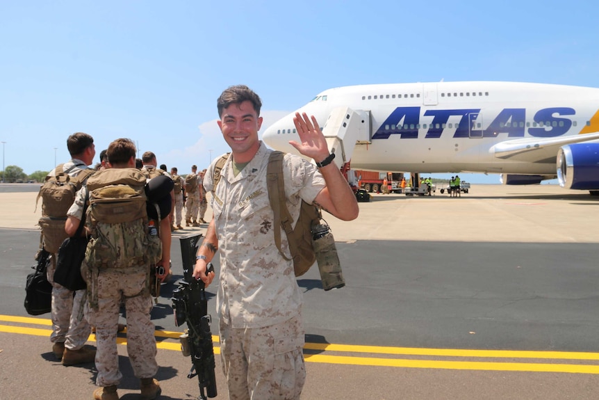 A US marine smiles and waves at the camera while lining up on the tarmac to board a plane.