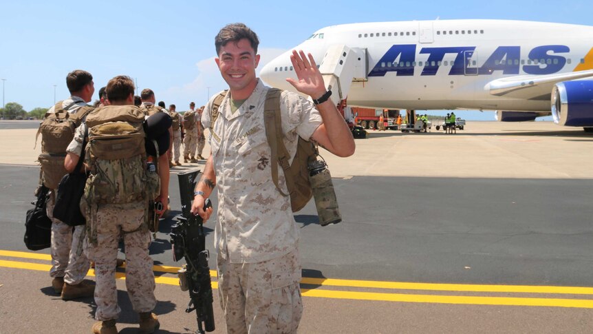 A US marine smiles and waves at the camera while lining up on the tarmac to board a plane.