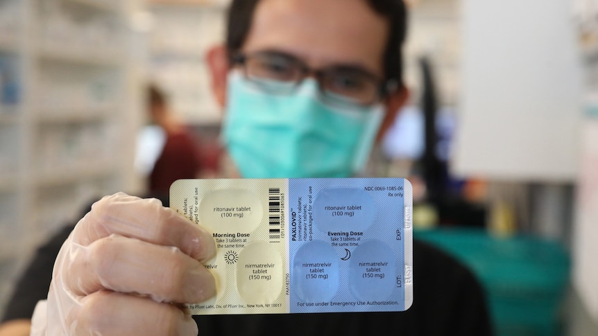 A man wearing a surgical mask holding up a packet of antiviral drugs