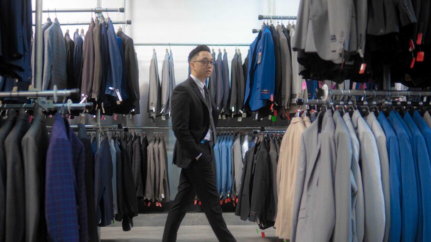 Man with sharp haircut, glasses and hands in pocked walks past racks of suits