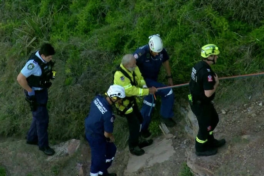 An elderly man still attached to paragliding ropes is escorted by emergency services.