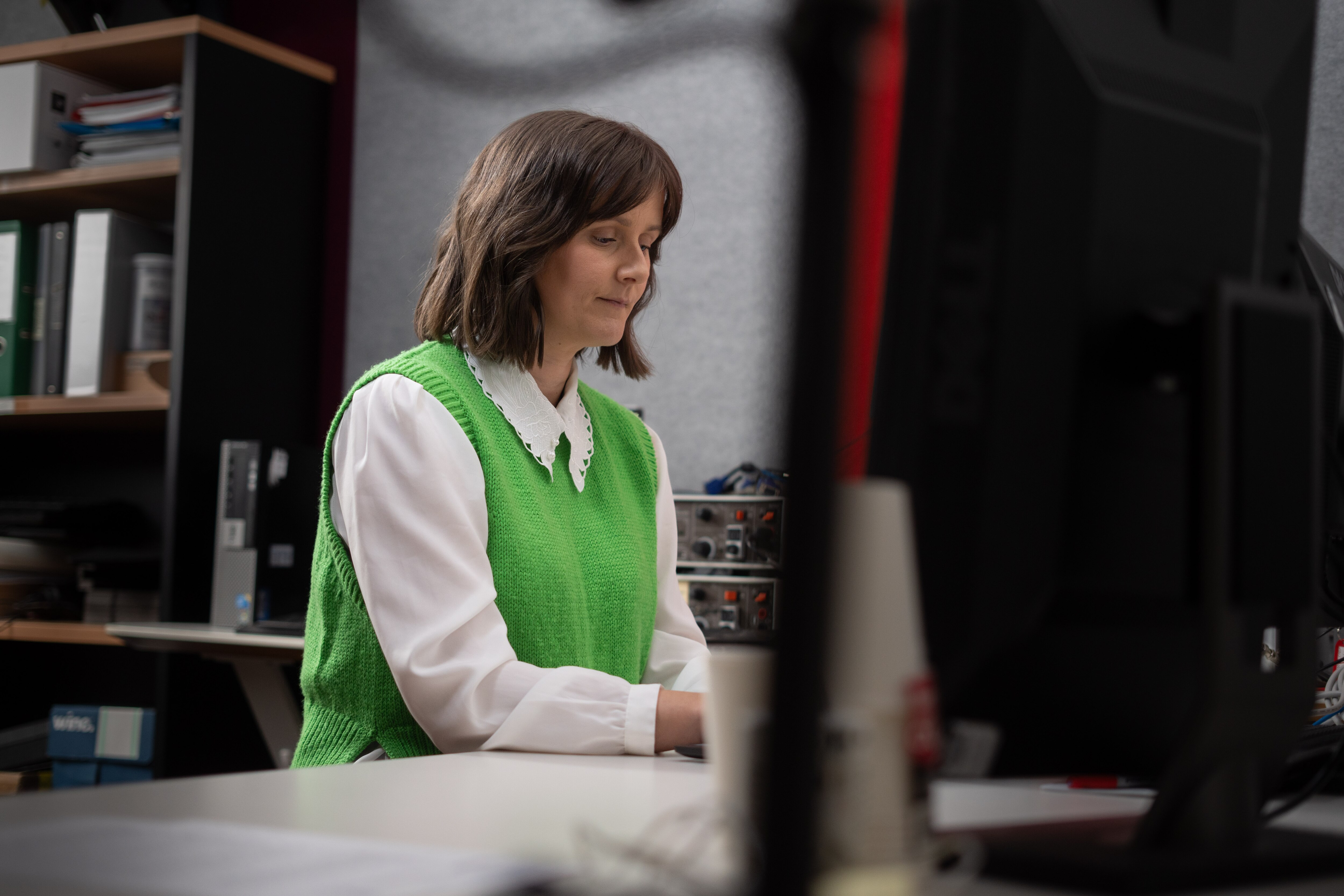 a woman sits at her computer in a green sweater vest and white shirt