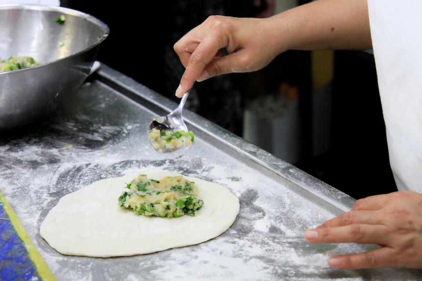 A hand holding a spoon hovers over food being prepared in a kitchen