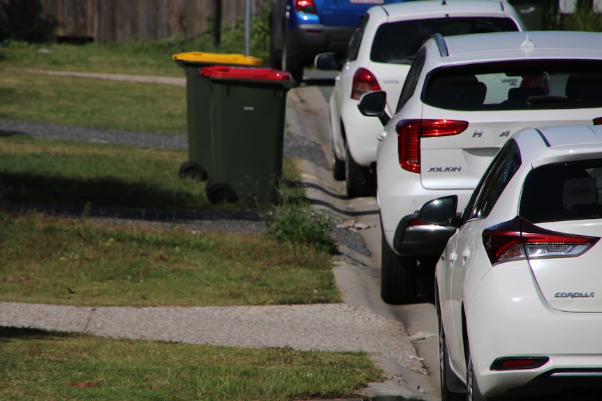The backs of three white cars parked on the left-hand side of the street with two council bins in the background