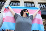 Person wearing rainbow face mask is seen from below looking up, holding outstretched trans flag, striped blue, pink and white.