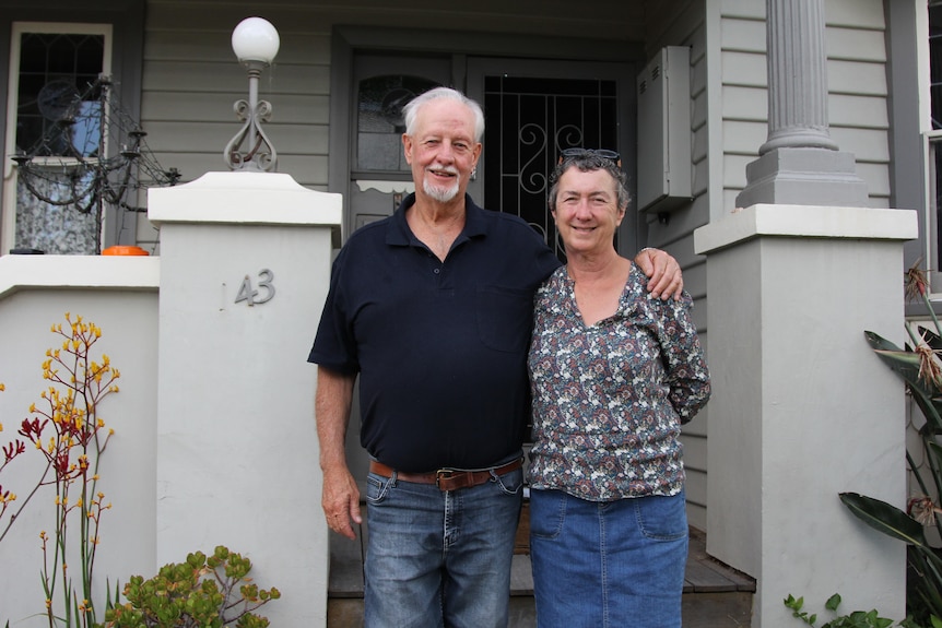 An older man and woman smiling at the camera in front of a house.