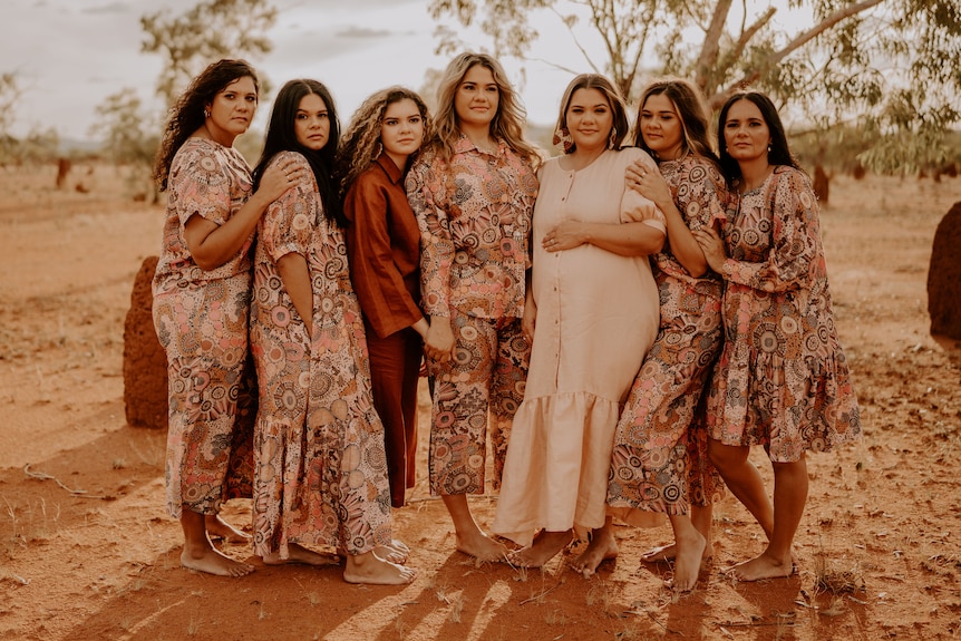 Seven Indigenous women pose for photo in desert landscape wearing colourful dresses.