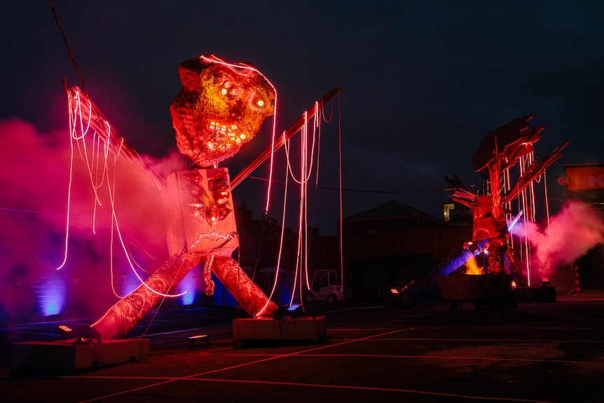 In a carpark at night, two enormous sculptures of fantastical creatures covered with orange and pink lights