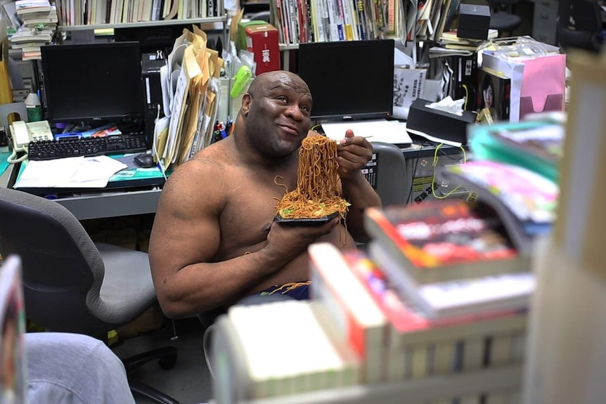 Bob Sapp sits in a crowded office without a shirt eating a plate of noodles.