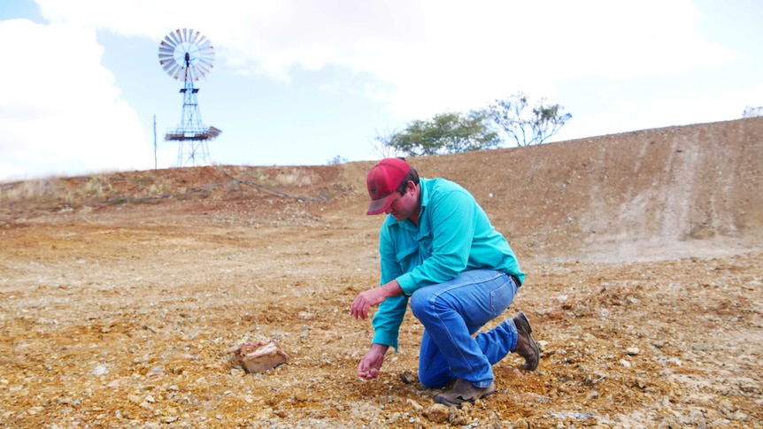 A man wearing boots blue jeans, an aqua shirt and red cap kneels down in the dirt.