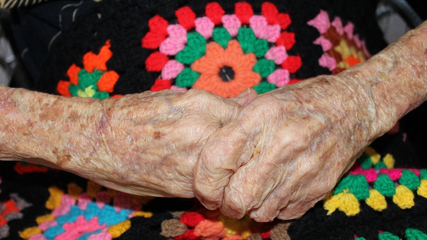 An elderly person's hands, clasped together over a colourful knitted blanket.