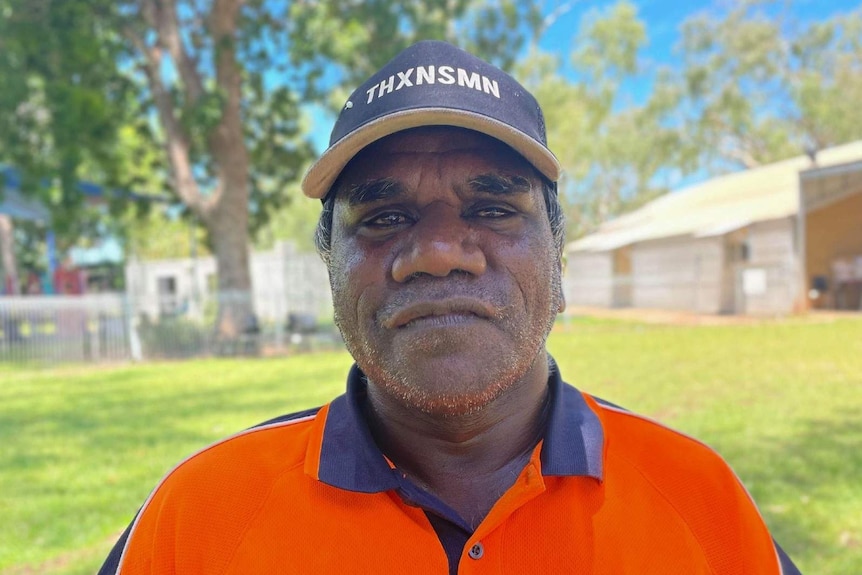 An Indigenous man in a cap and bright orange shirt stands and looks at the camera.