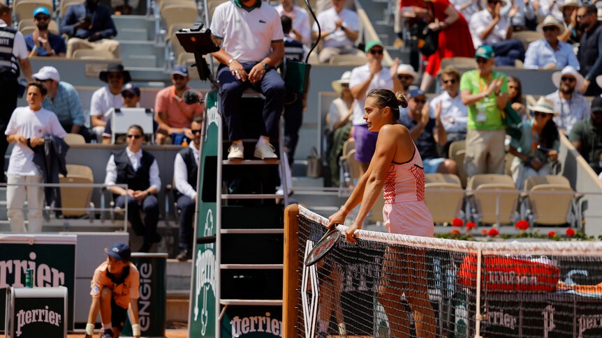 Tennis player waits at net on clay court.
