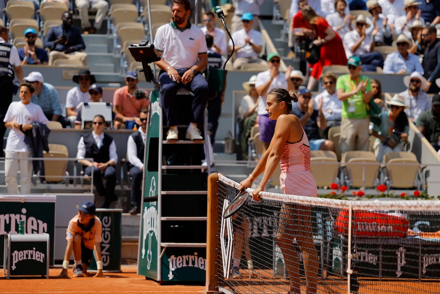 Tennis player waits at net on clay court.