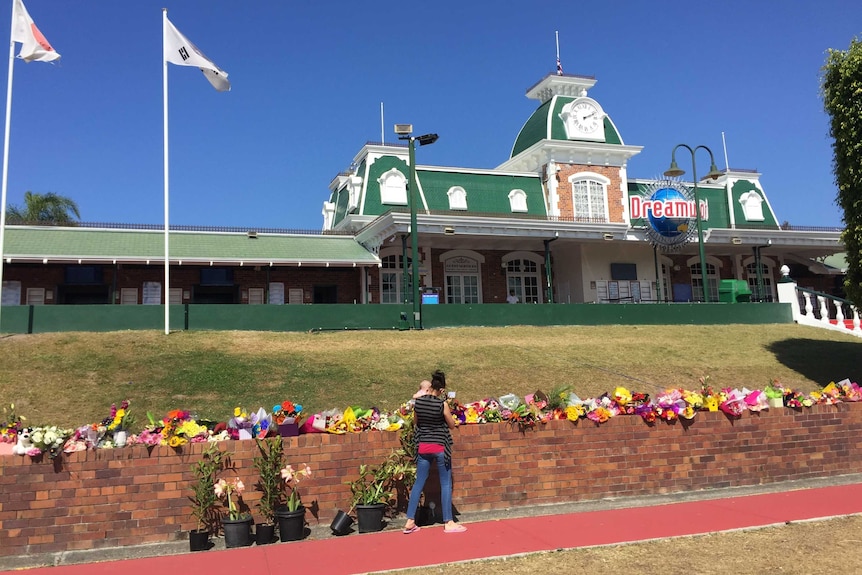 A woman holding a baby looking at floral tributes outside Dreamworld