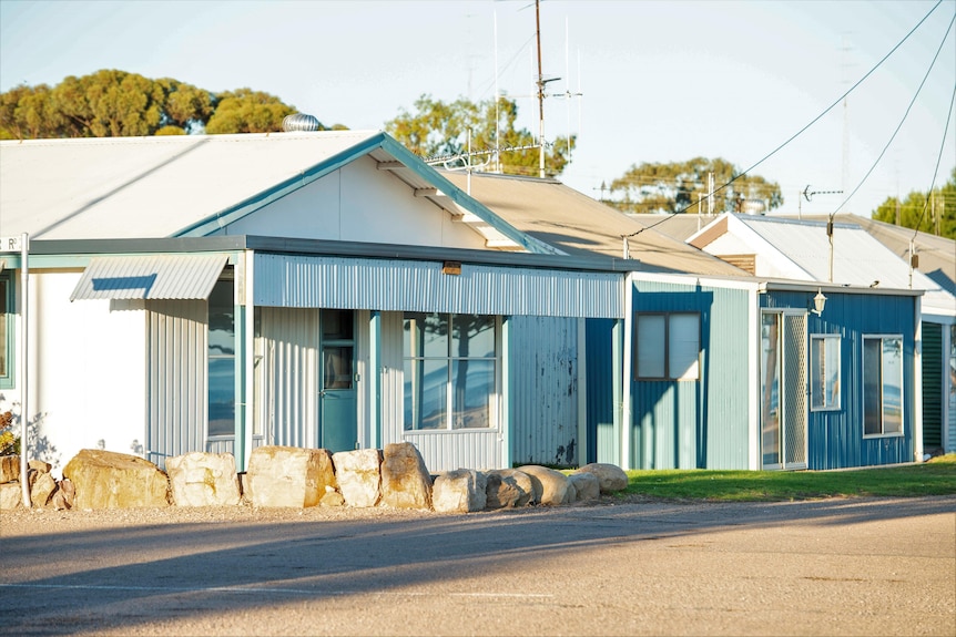 A collection of the front of several white and blue beach shacks against an unsealed road with TV antennas in the background.