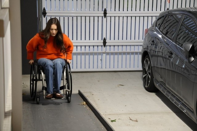 A young woman, dressed in jeans and an orange jumper, pushes her wheelchair up a ramp.