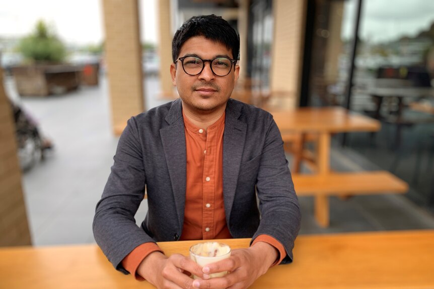 Shashi Karunanethy seated at a wooden bench, holding a coffee.