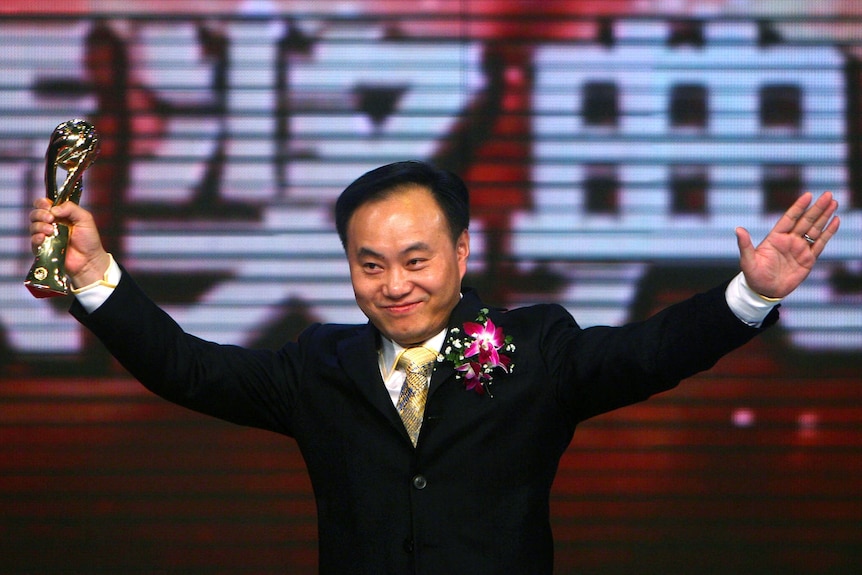 A man in a suit holds a golden trophy with both arms raised