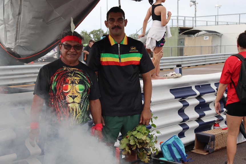 Two indigenous men pose for a picture, the younger one is holding a branch, with a cloud of smoke in the bottom left corner