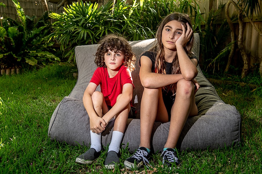Siblings Coby (l) aged nine and Maddy (r) aged 12 sit next to each other on a beanbag in their backyard looking bored.