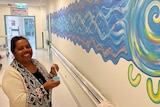 Woman on right smiling at camera with paint pot and brush next to blue mural on a long wall