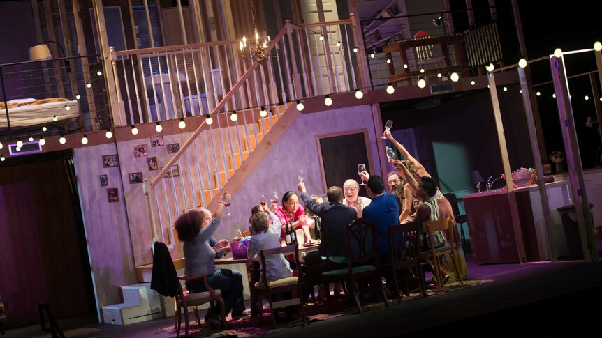 Production still showing a stage with an elaborate set for interior of home, and many people seated at a kitchen table.