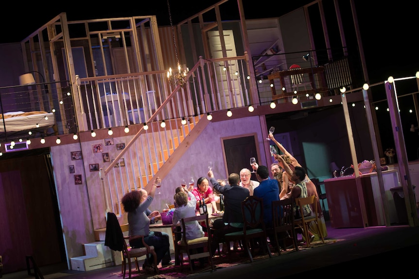 Production still showing a stage with an elaborate set for interior of home, and many people seated at a kitchen table.