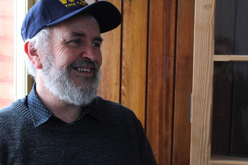 A man with grey hair and a beard, wearing a grey jumper and a blue cap.