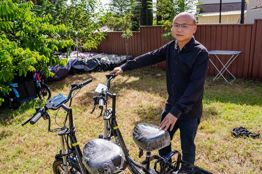 A man in a black shirt and glasses stands in a backyard with e-bikes, some under tarp and some not.