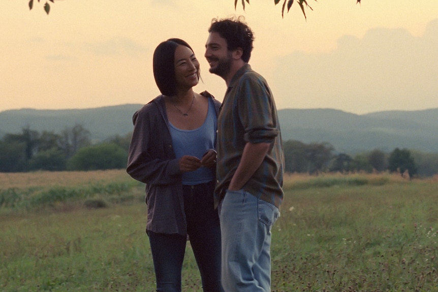 A Korean woman in pale blue top and dark jacket smiles at a white man in grey shirt and jeans, standing in a field at twilight.