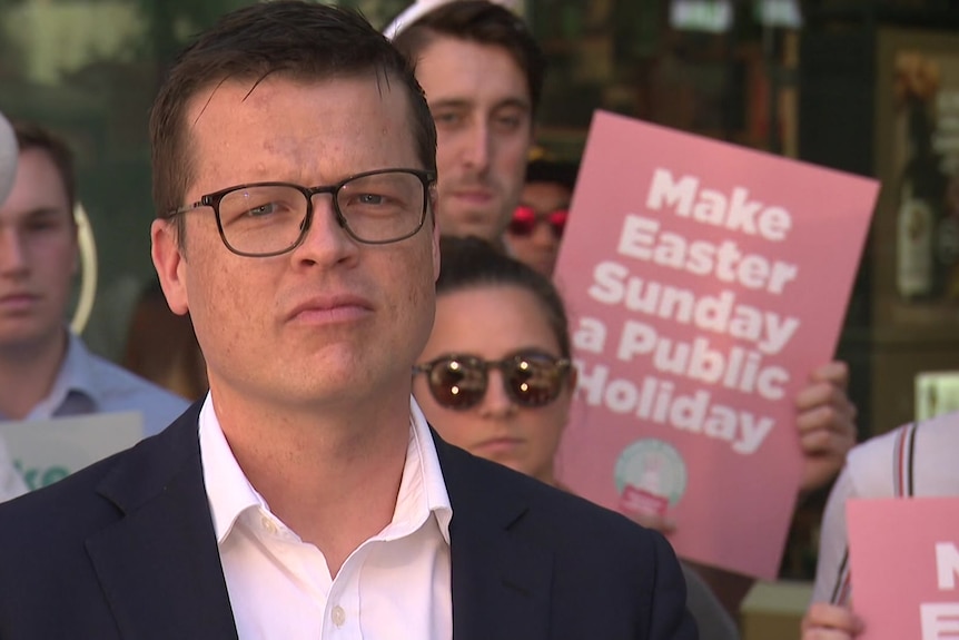 A man wearing a suit and glasses with people behind him holding pink posters reading 'make easter sunday a public holiday'