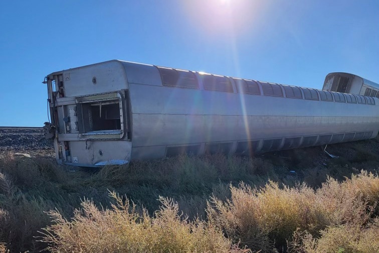 A carriage lies entirely on its side beside the tracks, having tipped over in the derailment.