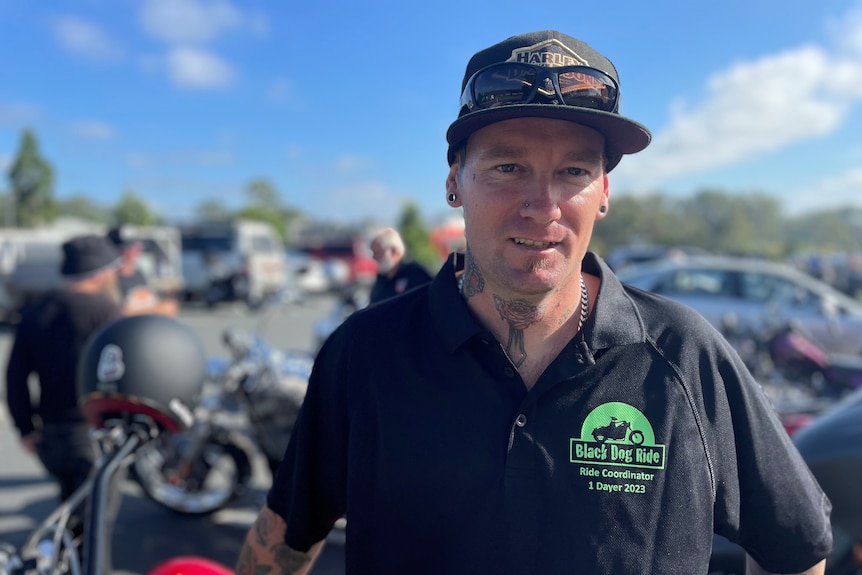 A man wearing a black t-shirt and black cap stands in front of motorbikes.
