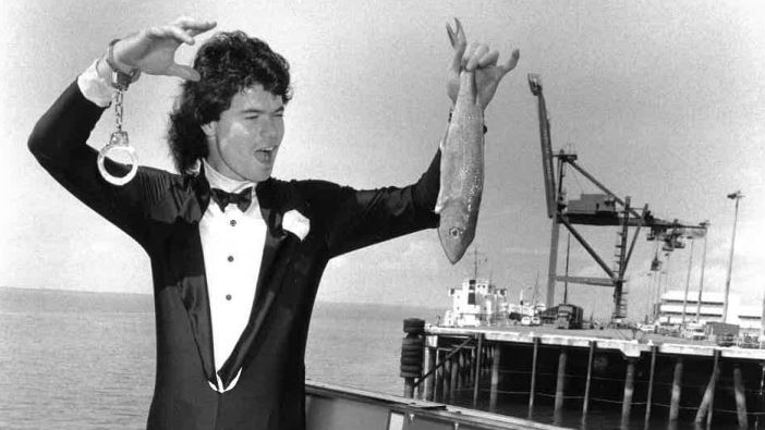 Tim hayward dressed in a tuxedo wears handcuffs and holds a fish